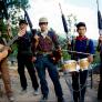 A group of five musicians on a rural road holding their instruments and rifles.